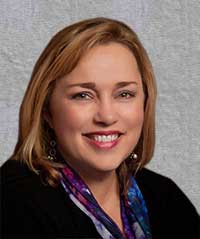 A woman with light brown hair smiles at the camera. She is wearing a black top and a colorful scarf with shades of blue and purple, embodying the professional yet approachable demeanor often seen in medical administrators. The background is a neutral gray tone.