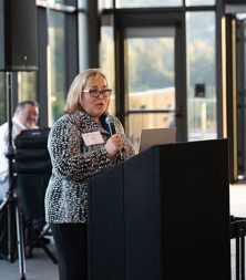 Jill Arena speaking at a podium with a microphone in a modern glass-walled venue. She is wearing glasses and a patterned jacket. There's a man sitting in the background, partially visible.